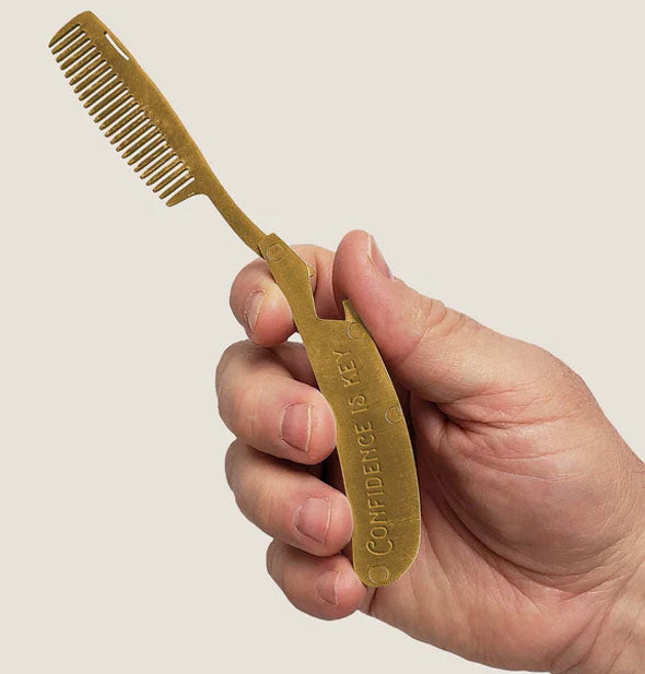 Model's hand holds a Confidence Is Key brass beard comb by its handle