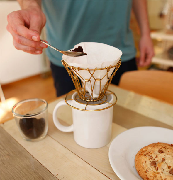 Model demonstrates use of the Brass Collapsible Coffee Dripper over a white mug on a wooden tabletop