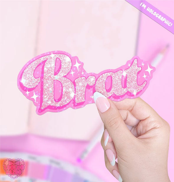 Model's hand holds a pink sticker that says, "Brat" in sparkly pink lettering accented by white stars