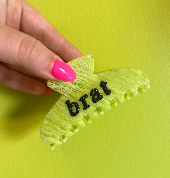 Model's hand holds a lime green hair clip with streaked effect and the word, "brat" in black accented with black rhinestones against a lime green backdrop