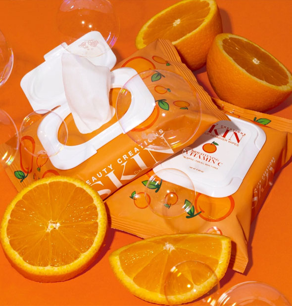 One closed and one opened pack of Brightening Vitamin C Beauty Creations Skin Makeup Remover Wipes staged with orange pieces and bubbles on an orange backdrop