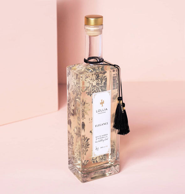 Tall, square, clear bottle of Lollia Elegance Bubble Bath with gold cap, floral patterning, and black tassels hanging from the neck against a pink backdrop