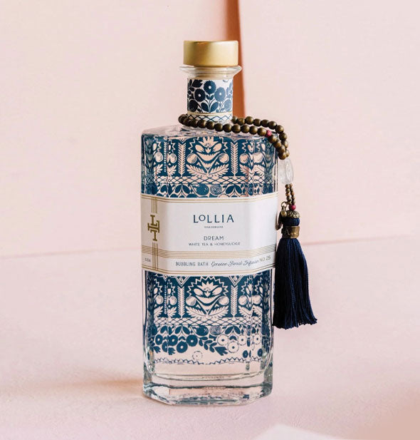 Glass bottle of Lollia Dream Bubble Bath features intricate blue patterning, a gold cap, and dark tassel on a beaded string hanging from its neck