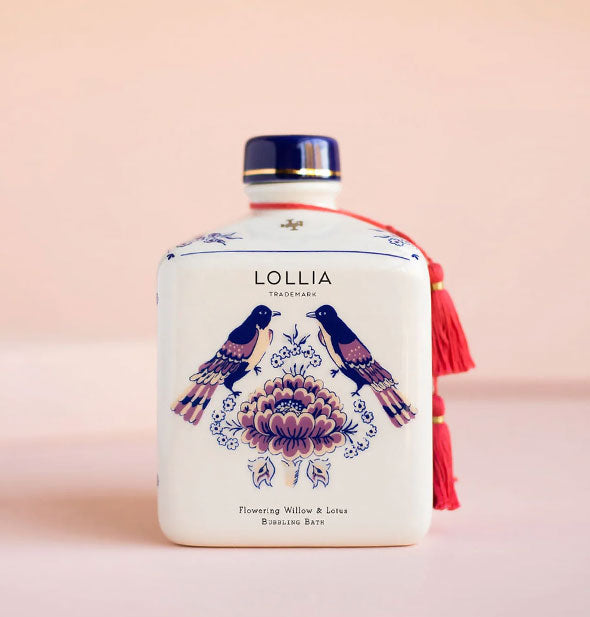 Square white Lollia Flowering Willow & Lotus Bubbling Bath bottle features a blue and purple design of birds and florals, a dark blue cap with gold accent stripe, and red tassels hung from its neck