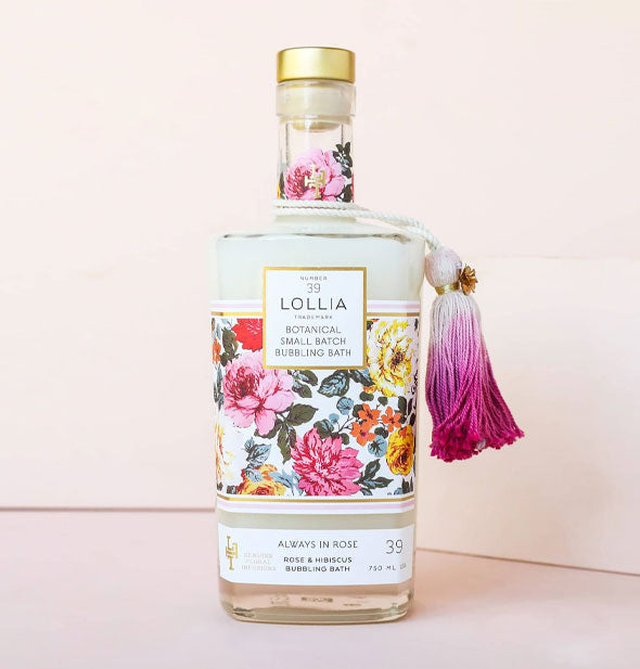 Glass bottle of Lollia Always In Rose Botanical Small Batch Bubbling Bath with colorful floral label, gold cap, and pink-to-white ombre tassel hanging from the neck