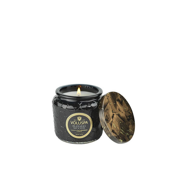 A lit Voluspa Burning Woods candle in patterned black glass jar with decorative tin lid alongside