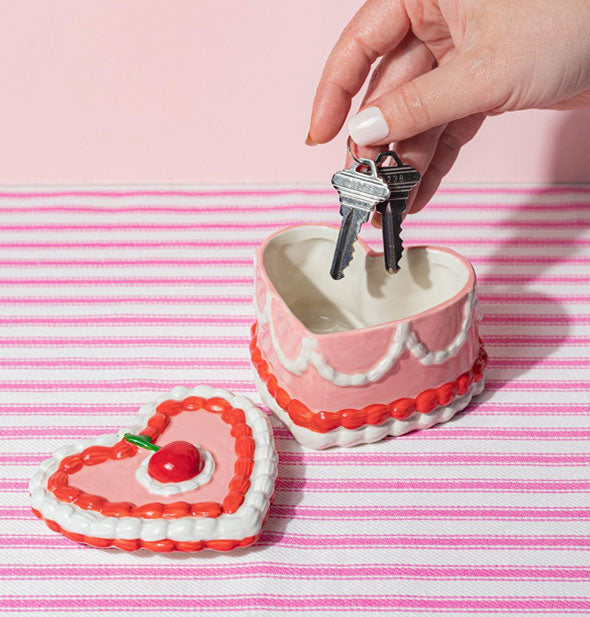 Model's hand places house keys into a ceramic heart-shaped cake box with lid resting on a pink striped surface