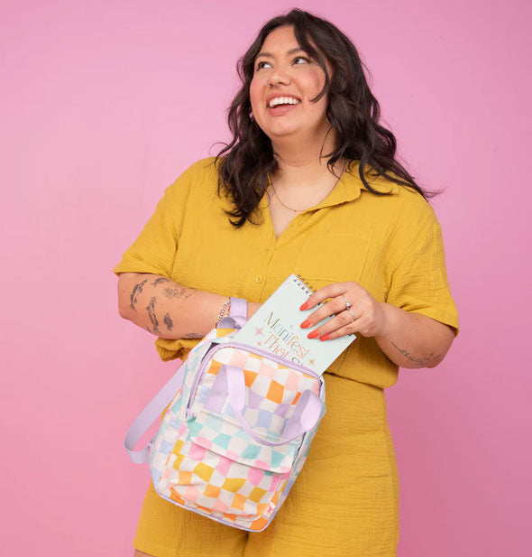 Smiling model puts a spiral-bound notebook into a Carnival Check Mini Backpack against a pink backdrop