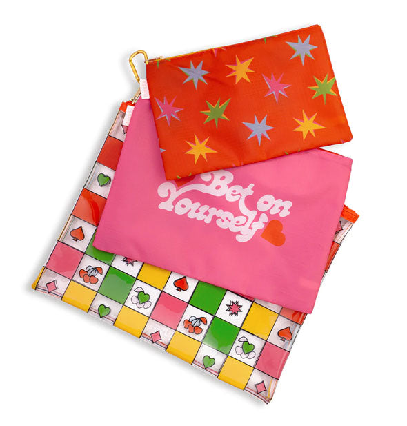 Set of three pouches joined together by a gold carabiner: Bottom pouch is multicolored checker print with various symbols, middle pouch is pink with, "Bet on Yourself" printed in white script with red heart, and top pouch is red with multicolor starburst pattern