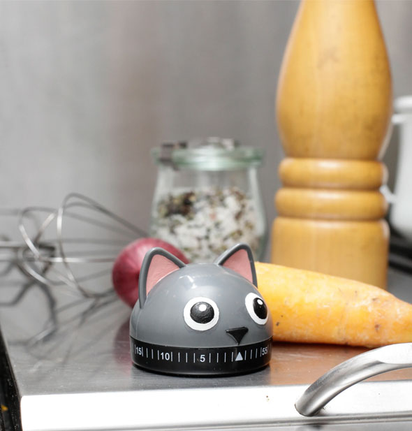 Gray cat head kitchen timer rests on a countertop with vegetables, utensils, and spices