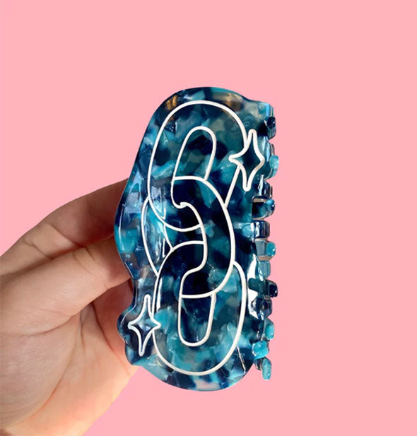 Model's hand holds a blue tortoise hair claw clip that features a white chain design with star accents in front of a pink backdrop