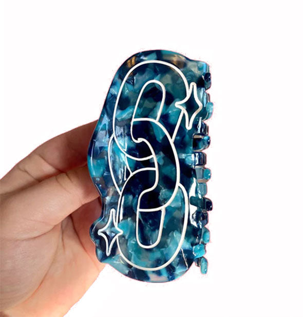 Model's hand holds a blue tortoise hair claw clip that features a white chain design with star accents