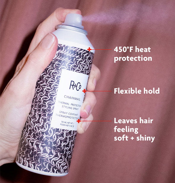 Model's hand dispenses a fine mist of R+Co Chainmail Thermal Protection Styling Spray from bottle against a mauve curtain backdrop; labels read, "450°F heat protection; Flexible hold; Leaves hair feeling soft + shiny"