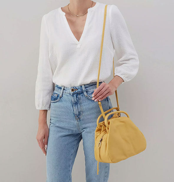 Model wearing jeans and a white shirt wears a pale yellow leather satchel bag over shoulder with an elongated strap