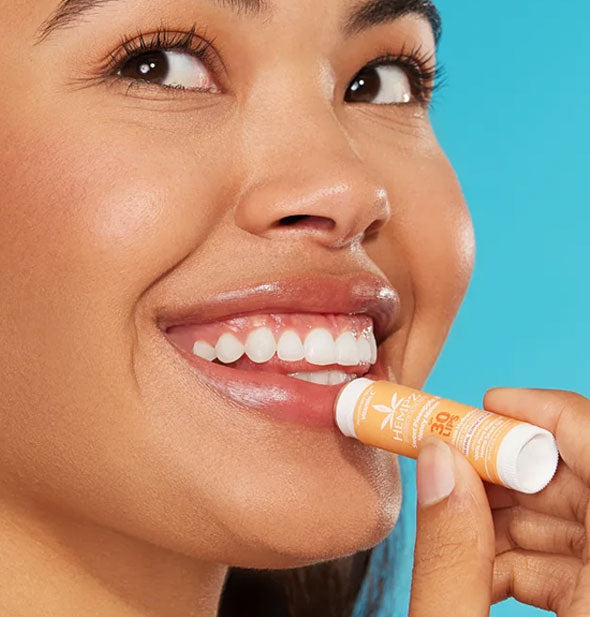 Smiling model applies Hempz lip sunscreen to bottom lip against a blue backdrop