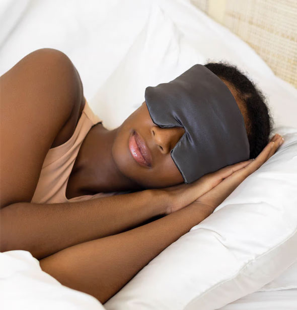 Model appearing to be sleeping on a white bedspread wears a charcoal satin eye mask