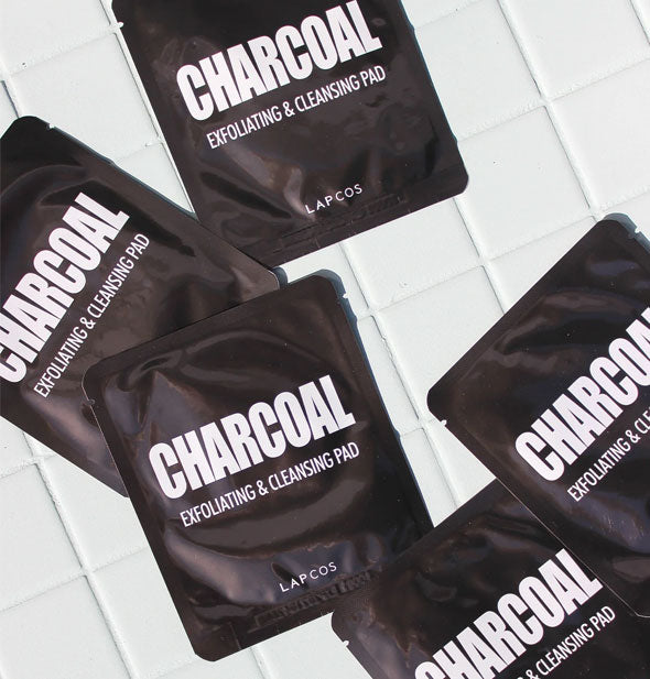 Assortment of black Lapcos Charcoal Exfoliating & Cleansing Pad packets on a white tiled surface