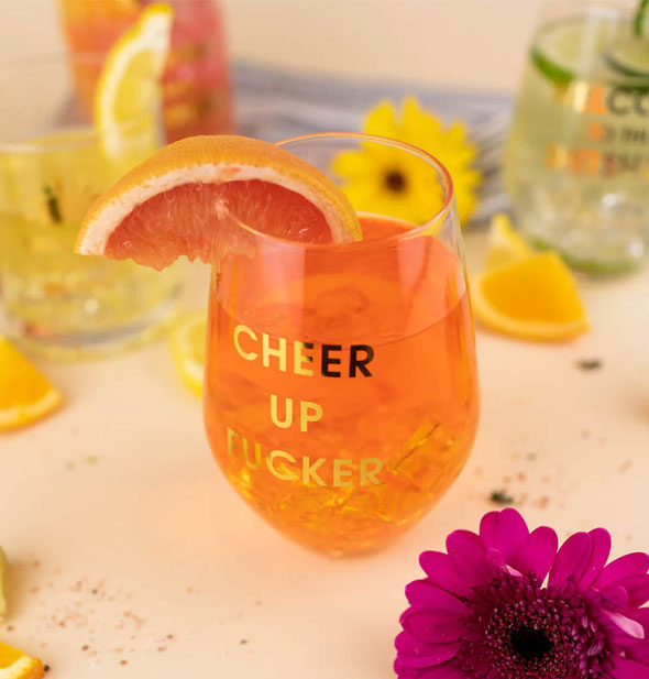 Cheer Up Fucker stemless wine glass holds an orange beverage and citrus slice garnish on a tabletop staged with flowers and other cocktail accouterments