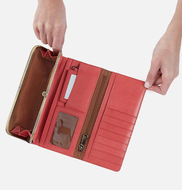 Model's hands hold open a salmon-colored leather trifold wallet with brass hardware and brown lining