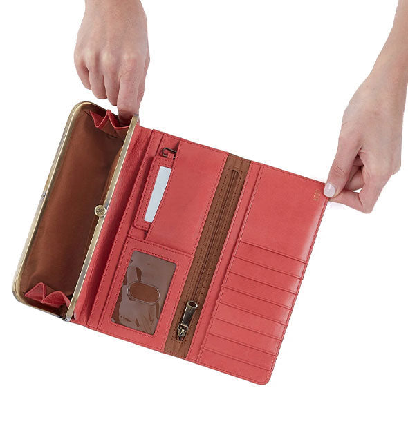 Model's hands hold open a salmon-colored leather trifold wallet with brass hardware and brown lining