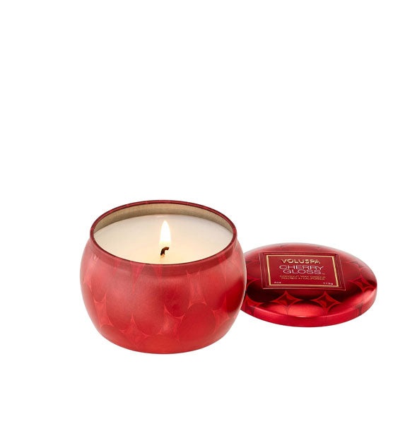 Lit candle in patterned metallic red tin vessel with lid set to the side