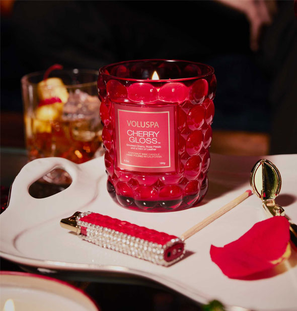 Lit Cherry Gloss Voluspa Candle on a white tray with rhinestone-encrusted lighter, matchstick, rose petal, and other items