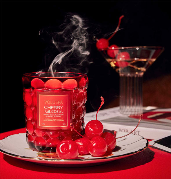 A Cherry Gloss Voluspa candle which has just had its flame extinguished smokes lightly on a dish beside a handful of cherries