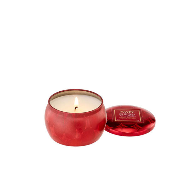 Lit candle in patterned metallic red tin vessel with lid set to the side