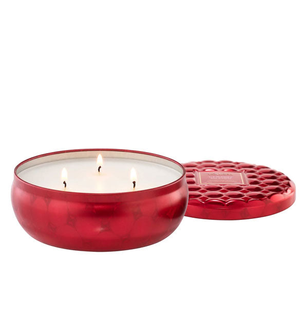 Lit three-wick candle in metallic red patterned tin vessel with textured lid set to the side and slightly behind