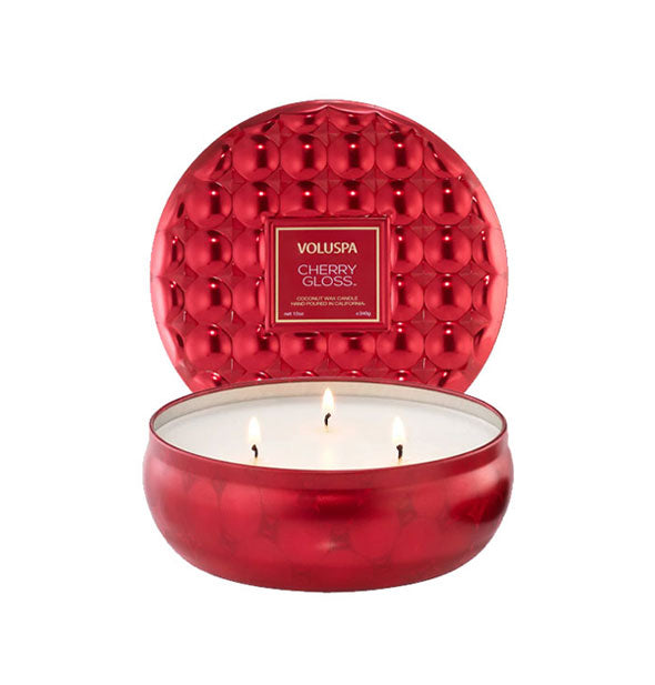 Lit three-wick candle in metallic red patterned tin vessel with textured lid set upright behind the tin