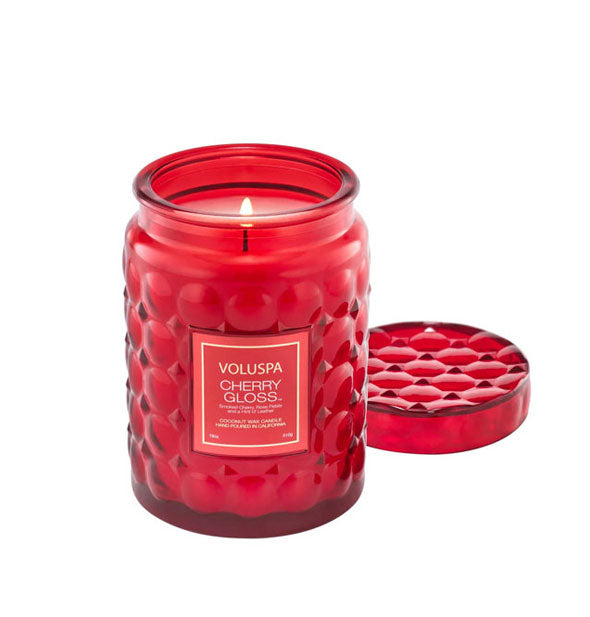 Lit Voluspa Cherry Gloss candle in a textured red jar with matching lid removed and set to the side and rear