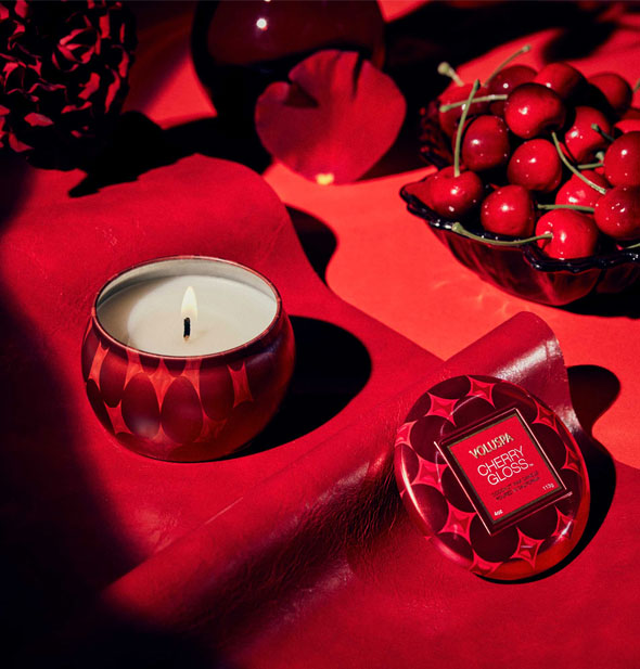 Lit Cherry Gloss tin candle on a piece of red leather with a bowl of cherries nearby