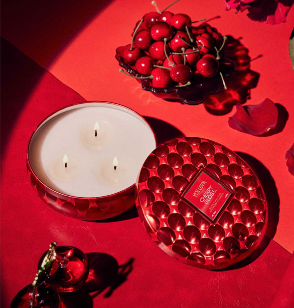 Lit three-wick candle in textured metallic red vessel on a red surface staged with cherries, red rose petals, and other items