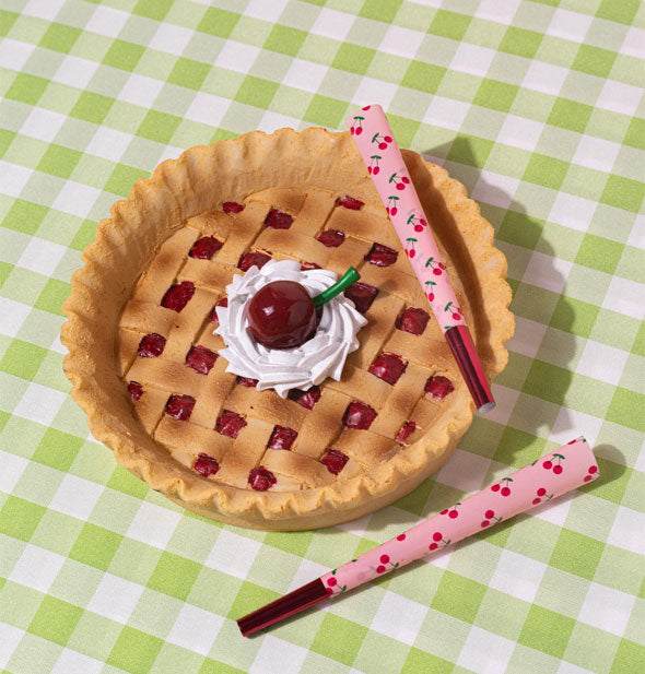 Round ashtray on a green gingham surface is designed to resemble a cherry pie with lattice crust is staged with two rolled cigarette with pink cherry-patterned paper