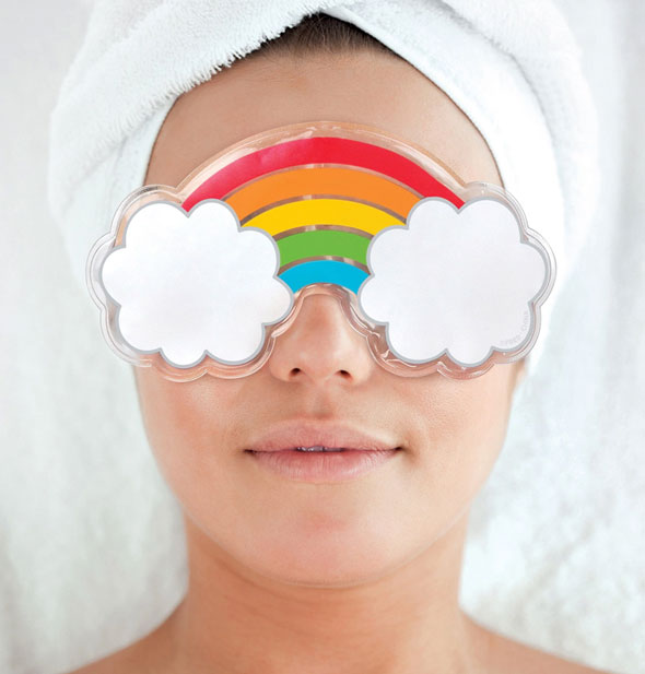 Model with hair wrapped up in a white towel wears a rainbow and white clouds cooling gel mask over eyes