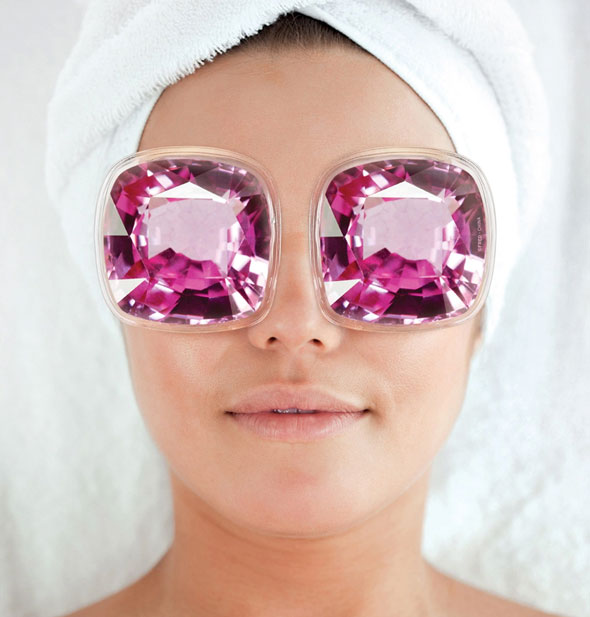 Model with hair wrapped up in a white towel wears a pair of purple gemstone cooling eye pads over eyes