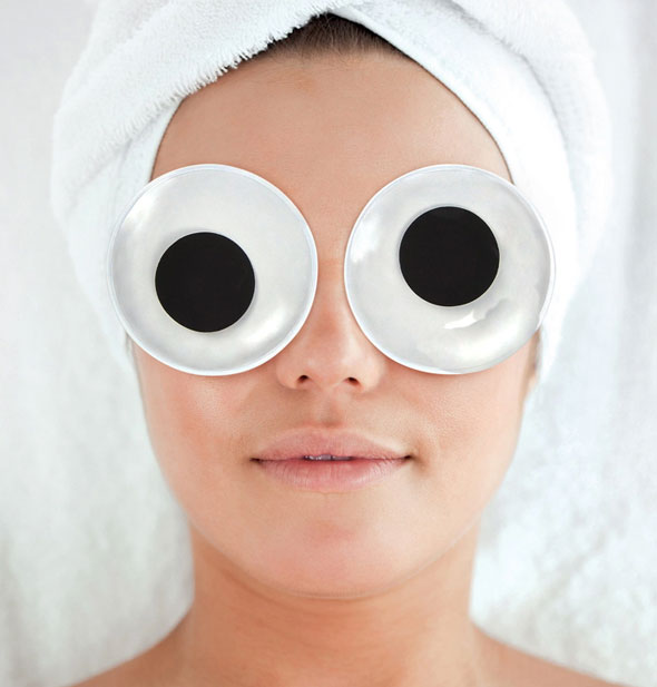 Model with hair wrapped up in a white towel wears a pair of googly eye cooling gel pads 
