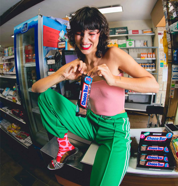 Model who appears to be struggling to unwrap a Choco Bar socks pack wrapper wears a sock of the same style with an open-toe heeled shoe propped on a convenience store counter