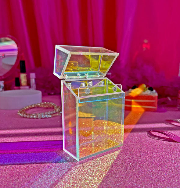 Clear acrylic cigarette box with iridescent prismatic finish rests on a sparkly pink surface with cosmetics and other items in the background