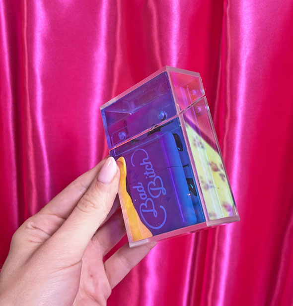Model's hand holds a prismatic acrylic cigarette box filled with Bad Bitch lighter and other items  in front of a pink satin curtain