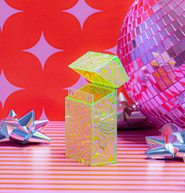 Neon green box with hinged lid and all-over psychedelic swirl pattern rests on a striped surface against a red and pink star background with holographic gift bows and a disco ball