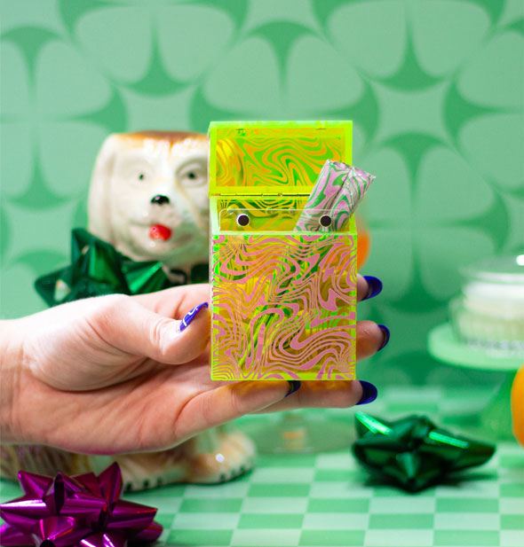 Model's hand holds a neon green swirl cigarette case with two rolled cigarettes inside against a green patterned background with gift bows and a ceramic dog figurine