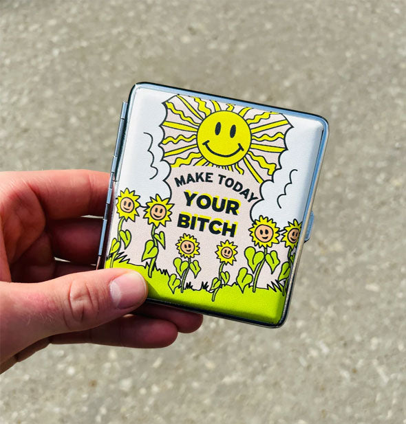 Model's hand holds a square case with illustration of a smiling sun peering out from between two puffy white clouds at a field of smiling sunflowers and the words, "Make today your bitch" in the center