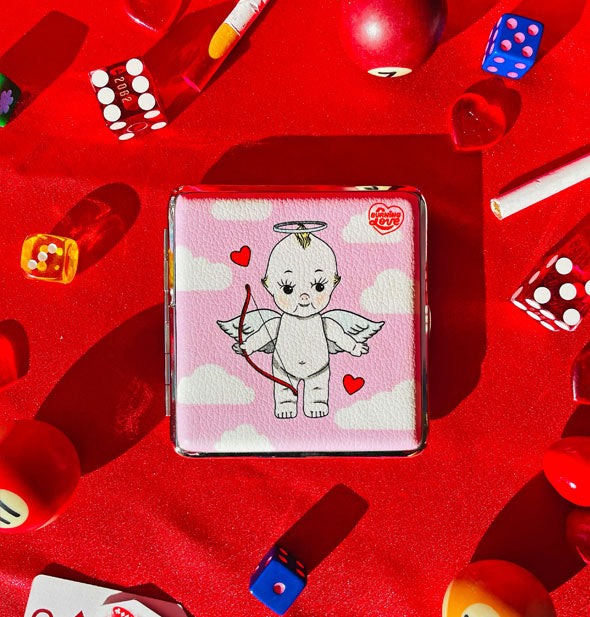 Square case on red surface scattered with dice, pool balls, and other items features Cupid Kewpie doll artwork