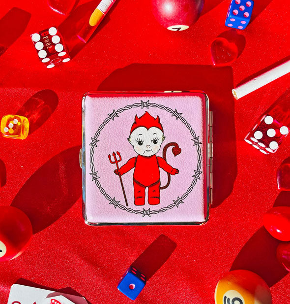 Square case on red surface scattered with dice, pool balls, and other items features devil Kewpie doll artwork
