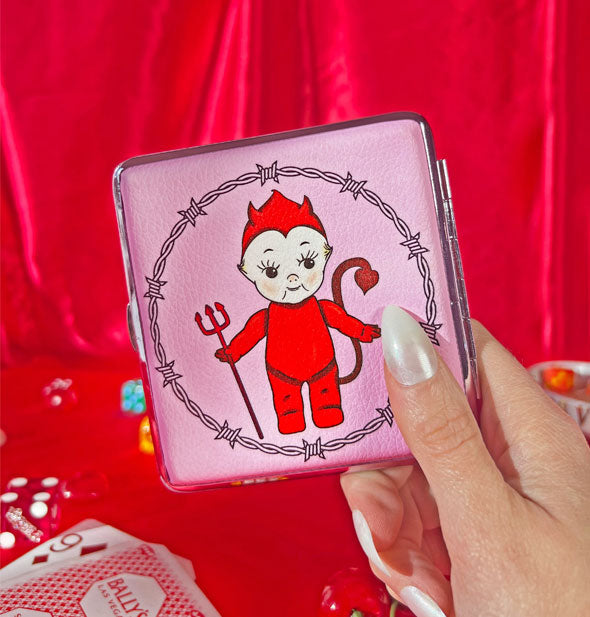 Model's hand holds a square devil Kewpie doll case with silver rim and hinge against a red backdrop scattered with game paraphernalia