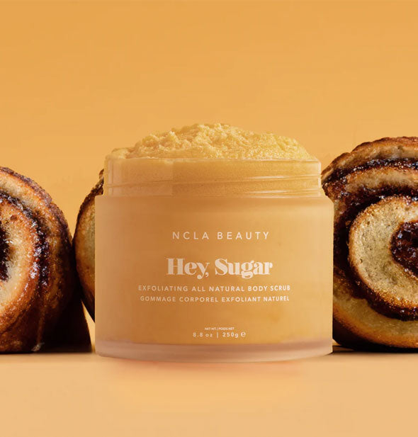 Opened tub of NCLA Beauty Hey, Sugar Exfoliating All Natural Body Scrub is surrounded by slices of cinnamon roll