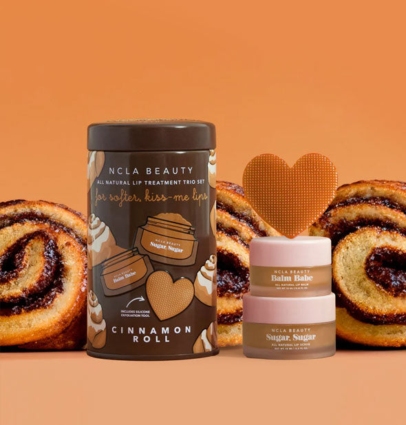 Contents of the Cinnamon Roll NCLA Beauty All Natural Lip Treatment set: Balm, scrub, and heart-shaped textured scrubber with tin surrounded by slices of cinnamon roll