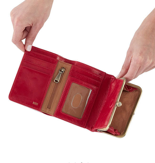 Model's hands hold open a red leather tri-fold wallet with brass hardware and brown lining