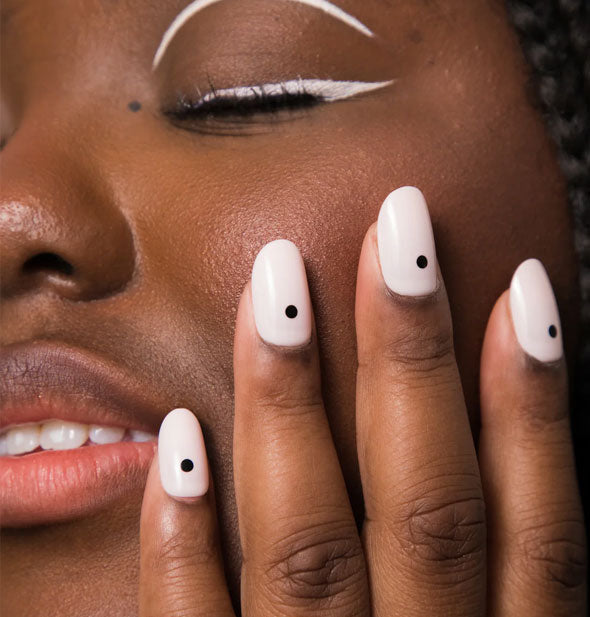 Model touches hand to cheek to show fingernails that are white with a single black dot on each
